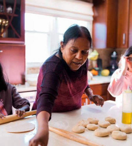 Two young Indian sisters stands at the kitchen counter with grandmother, learning to cook during a relaxing day at home together.